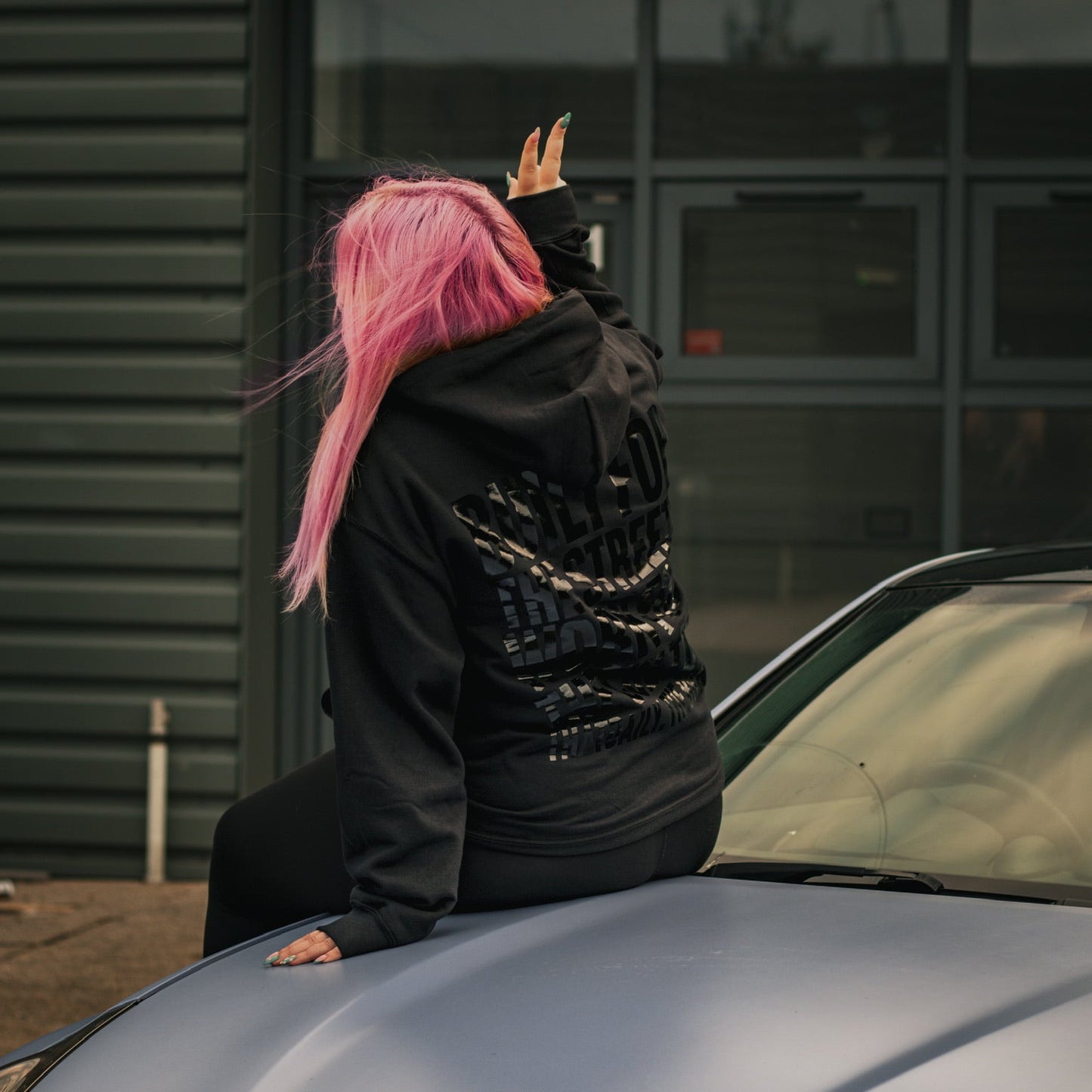 Person with pink hair and black hoodie on a car bonnet, waving.